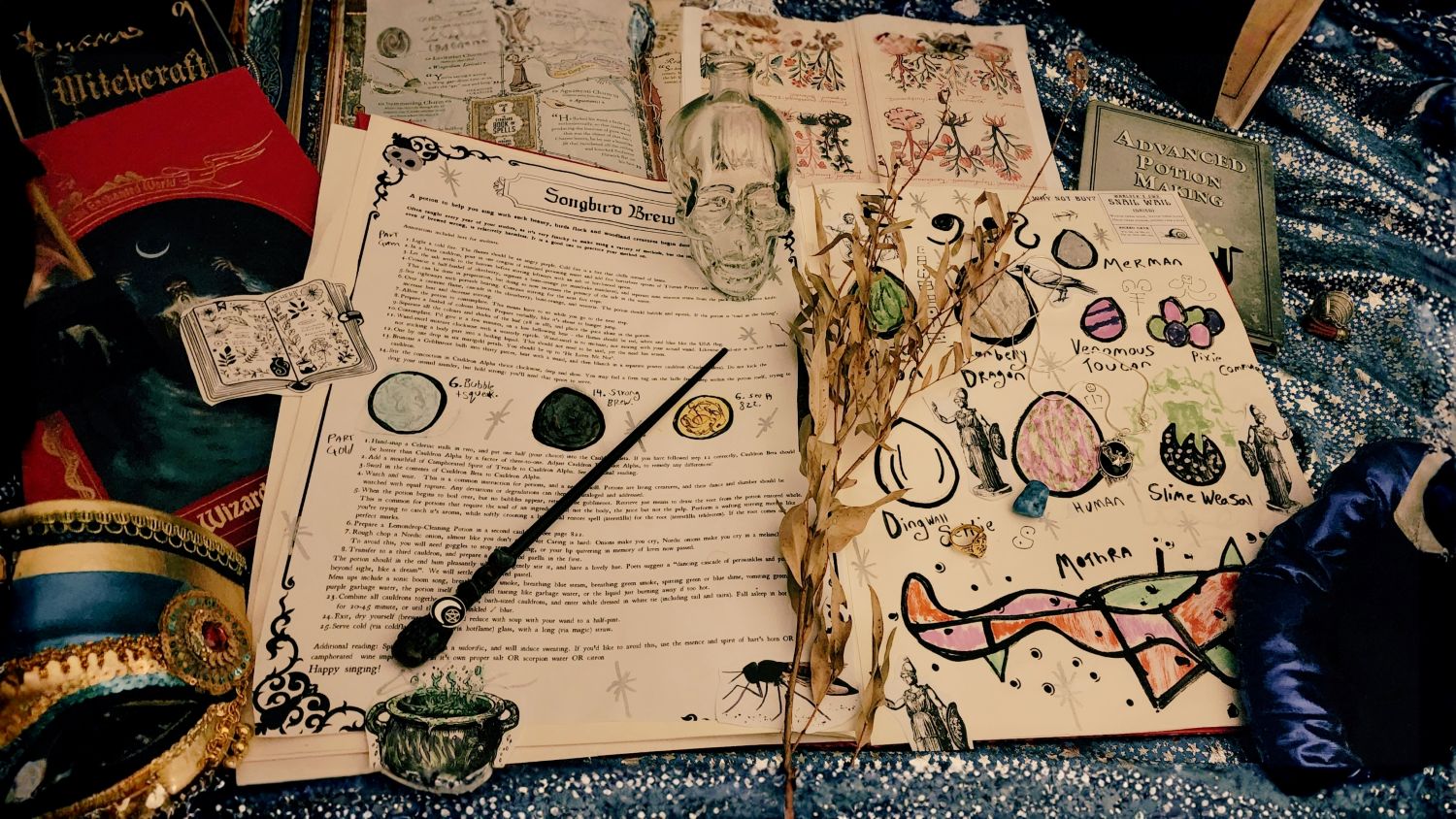 A collection of fantasy books, hand-drawn potion journals, a black magic wand, and a glass skull bottle are arranged over a blue, star-patterned background fabric.